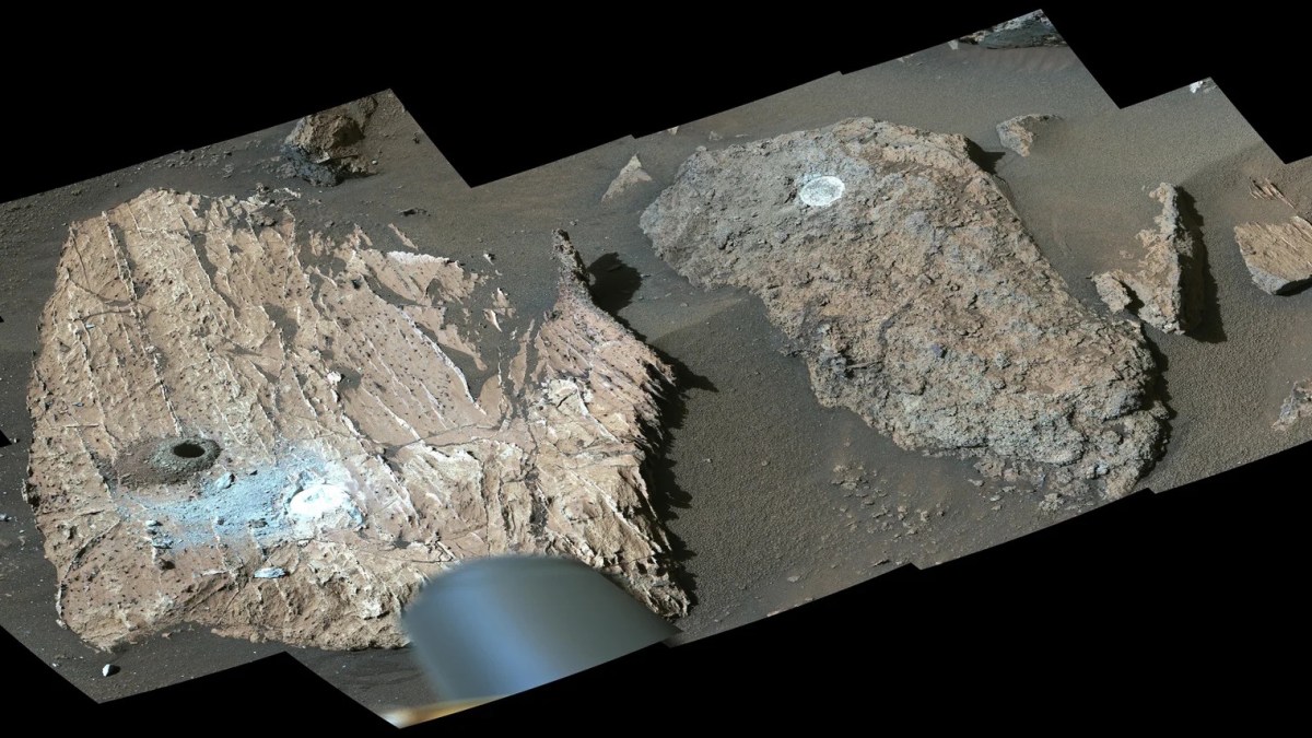 Two large Martian rocks with circular drill holes and light-colored dust are shown on a sandy, rocky surface. Part of a rover’s equipment, searching for traces of organics or signs of Mars life, is visible at the bottom of the image.