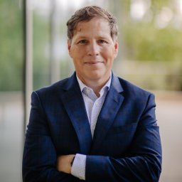 A man in a blue checked suit jacket and white shirt stands indoors with arms crossed, smiling softly at the camera. The background is out of focus with green and glass elements.