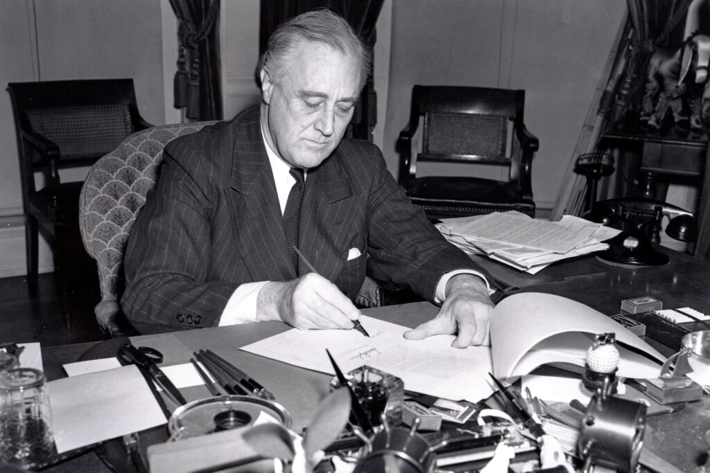 A man in a suit sits at a cluttered desk, signing documents with a pen. Papers, office supplies, and a telephone are visible on the desk.