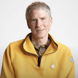 A man with short gray hair and a light complexion wears a yellow zip-up fleece over a plaid shirt, sitting against a plain white background.