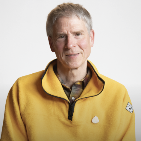 A man with short gray hair and a light complexion wears a yellow zip-up fleece over a plaid shirt, sitting against a plain white background.