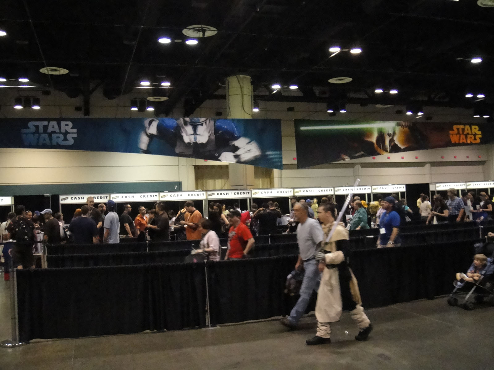 People walk through a convention hall with Star Wars banners overhead and queues for registration or ticket counters in the background.
