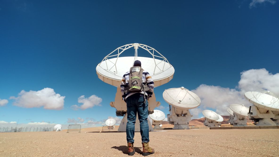 A person with a backpack stands in front of large satellite dishes in a desert area under a blue sky with scattered clouds.