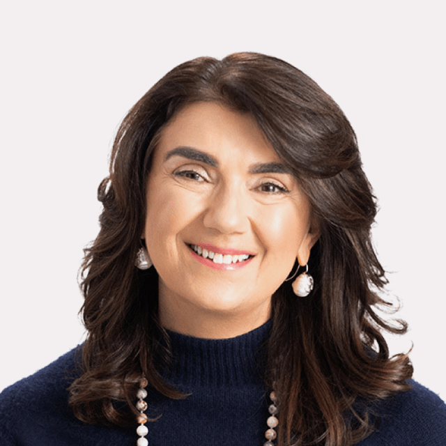 A woman with medium-length brown hair, wearing a navy sweater, pearl earrings, and a beaded necklace, smiling at the camera against a plain light background.