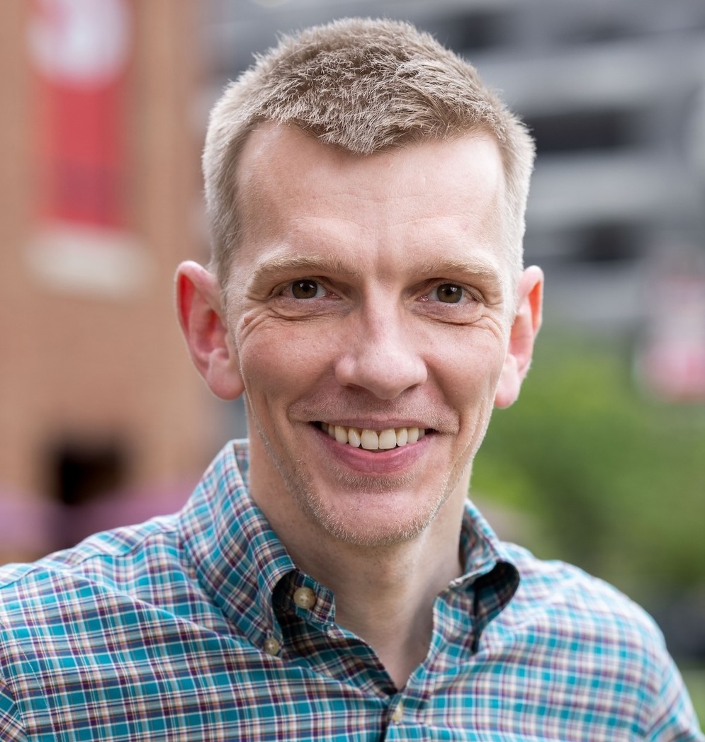 A man with short light brown hair wearing a plaid shirt smiles at the camera outdoors, with a blurred city background.