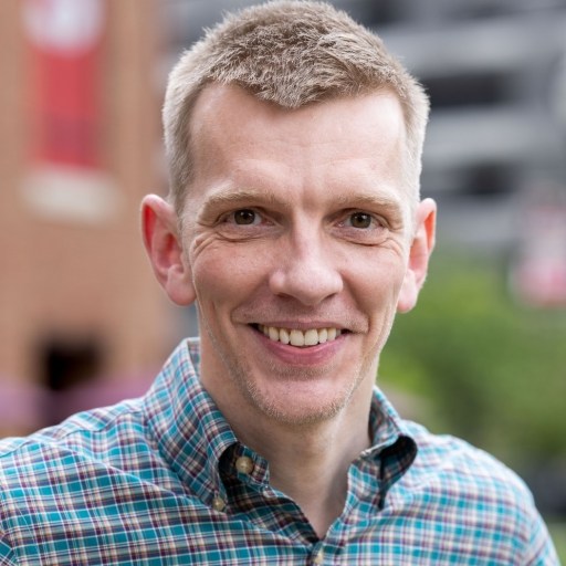 A man with short light brown hair wearing a plaid shirt smiles at the camera outdoors, with a blurred city background.