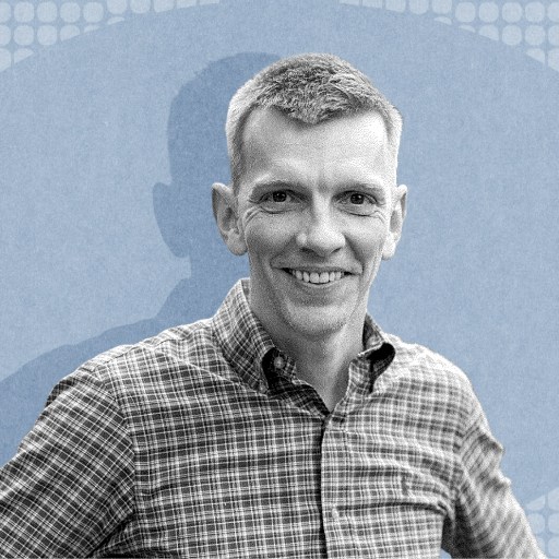 Angus Fletcher, wearing a plaid shirt, smiles at the camera as he stands in front of a blue, patterned background.