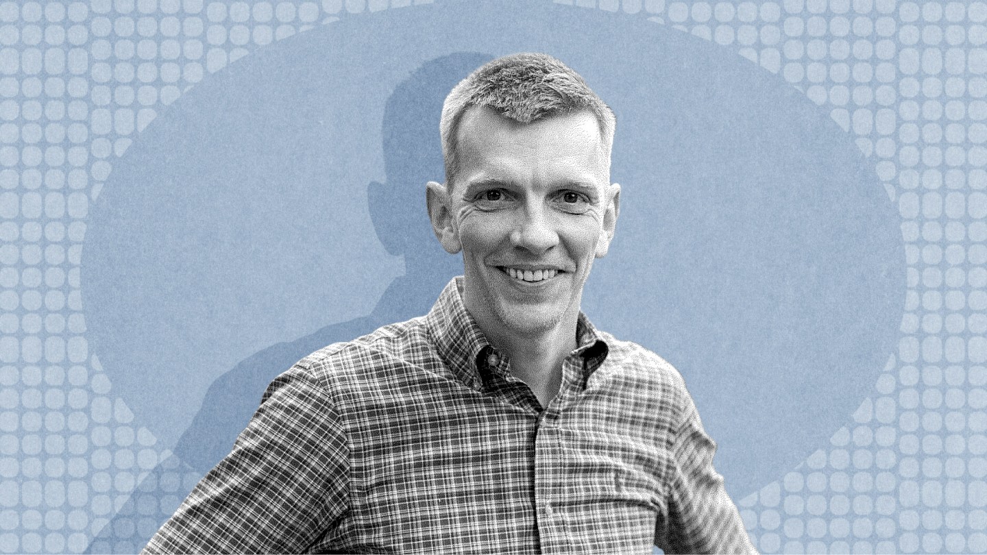 Angus Fletcher, wearing a plaid shirt, smiles at the camera as he stands in front of a blue, patterned background.