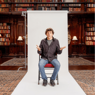 A man sits on a chair in front of a white backdrop, with bookshelves and lamps in the background, raising his hands in a questioning gesture.
