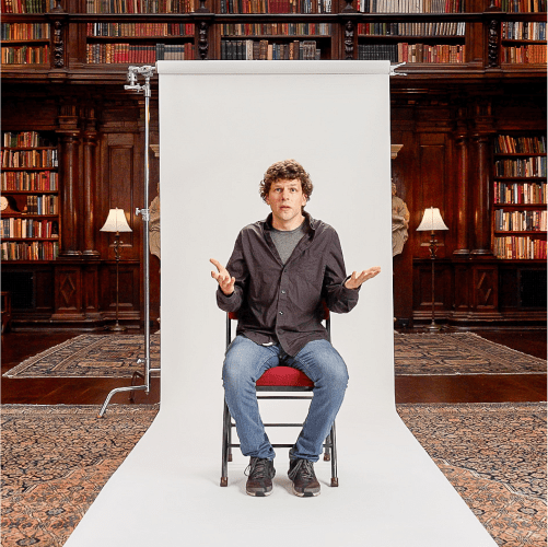 A man sits on a chair in front of a white backdrop, with bookshelves and lamps in the background, raising his hands in a questioning gesture.