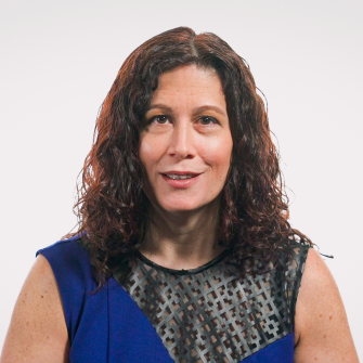 A woman with curly brown hair wearing a blue dress with a patterned black mesh panel, facing forward against a plain light background.
