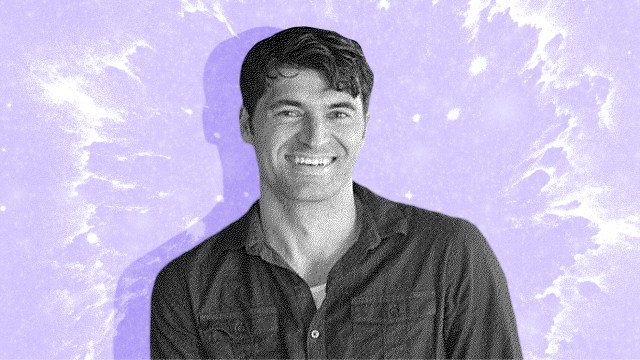 A smiling man with short dark hair wears a button-up shirt, standing in front of a purple, splattered-texture background.
