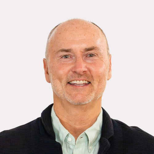 A middle-aged man with short light hair, a trimmed beard, and a light green shirt under a dark jacket, smiling at the camera against a plain background.