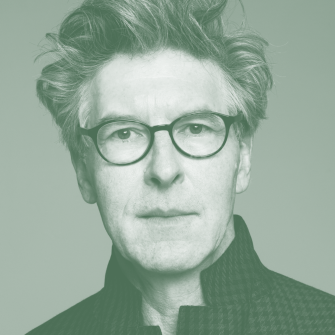 A man with tousled hair and glasses looks straight at the camera, wearing a high-collared, textured jacket. The image has a green tint.