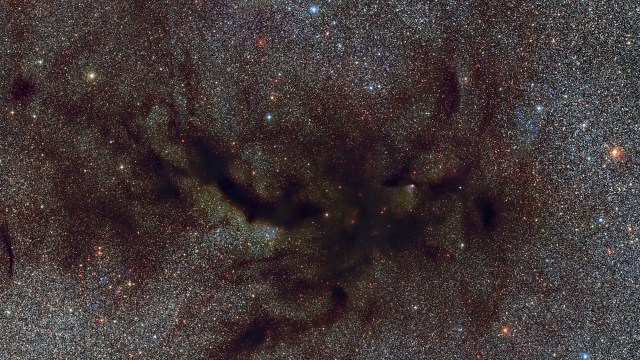 A dense star field with dark, irregular dust clouds—where cosmic dust come from—obscures parts of the glowing stars in the Milky Way.