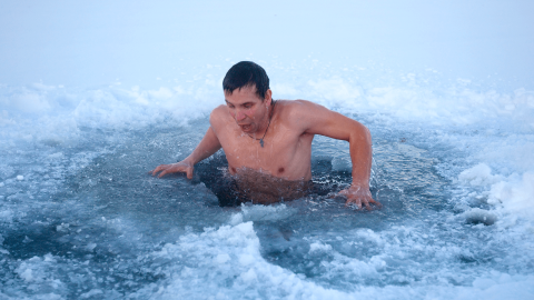 A man emerges from an ice hole in a frozen body of water, surrounded by snow and ice, taking a cold plunge during winter.