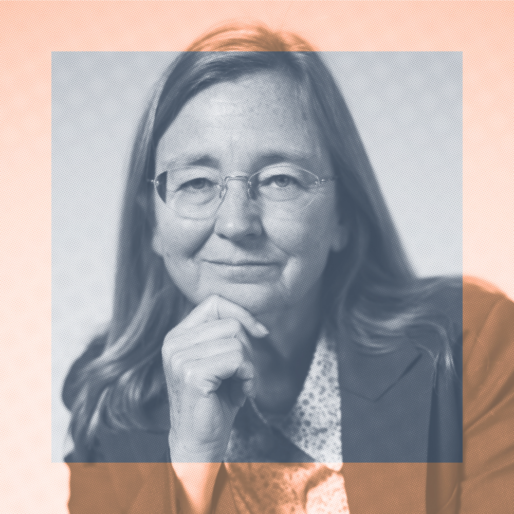 A person with long hair and glasses, wearing a patterned shirt and jacket, rests their chin on their hand and looks at the camera.