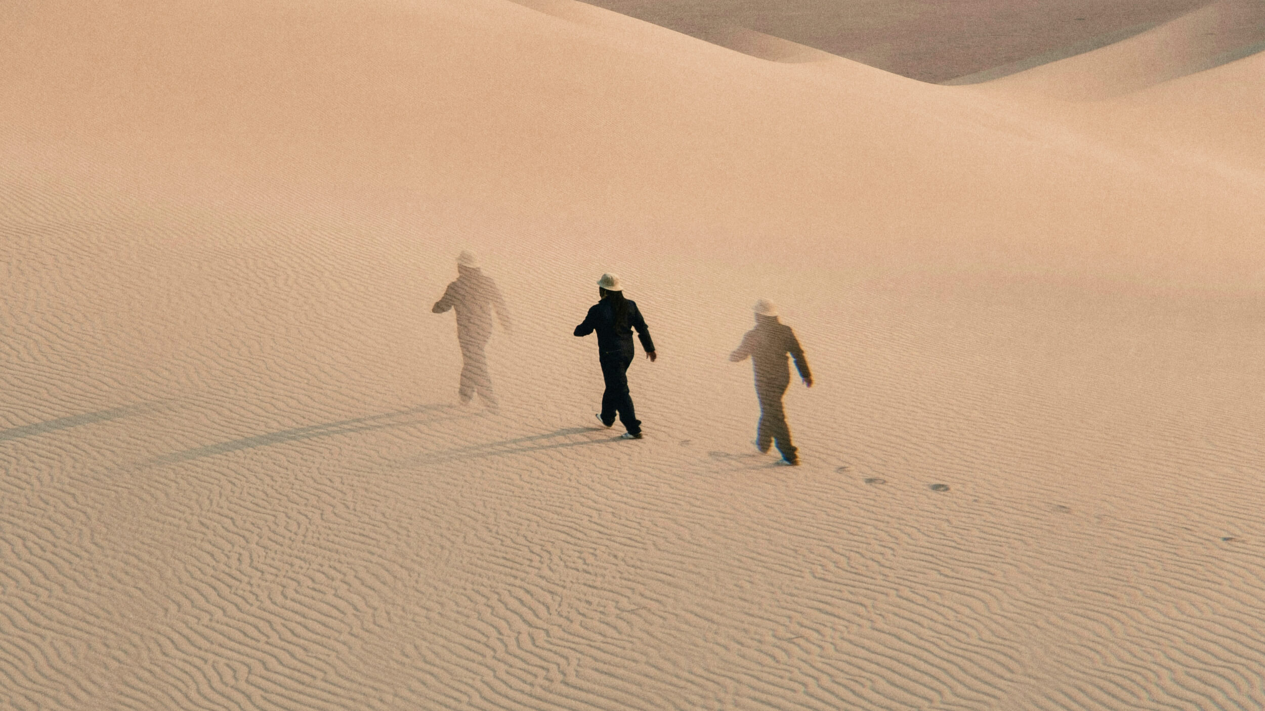 Three figures in hats walk across rippled sand dunes, leaving footprints behind them under soft, diffused light.