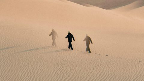 Three figures in hats walk across rippled sand dunes, leaving footprints behind them under soft, diffused light.