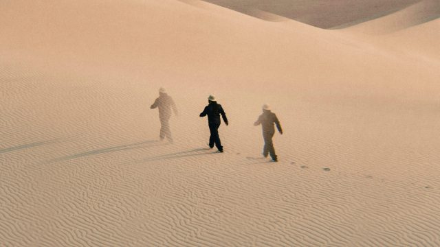 Three figures in hats walk across rippled sand dunes, leaving footprints behind them under soft, diffused light.
