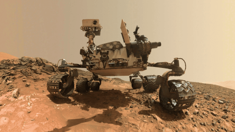 Mars rover on rocky terrain, showing its camera mast, equipment, and six wheels against a dusty, reddish Martian landscape.