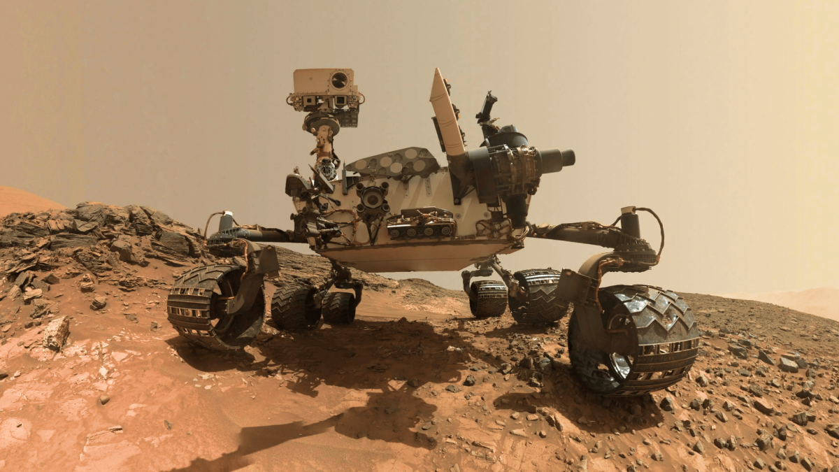 Mars rover on rocky terrain, showing its camera mast, equipment, and six wheels against a dusty, reddish Martian landscape.