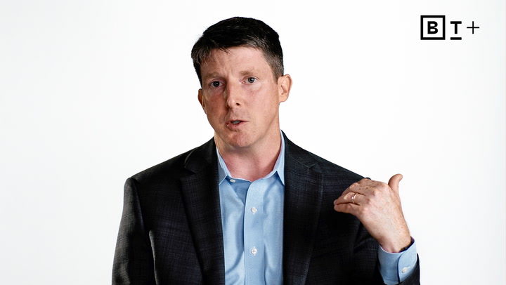 A man in a suit jacket and blue shirt gestures with his right hand, speaking, against a plain white background with a "BT+" logo in the top right corner.