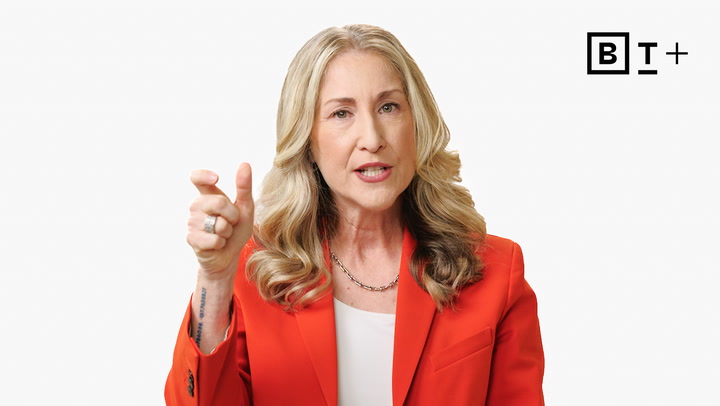 A woman with long blonde hair wearing a red blazer gestures with her hand while speaking against a plain white background. A "BT+" logo appears in the top right corner.