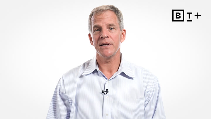 A man in a light blue button-down shirt speaks in front of a white background with a small microphone clipped to his collar. The "BT+" logo appears in the top right corner.