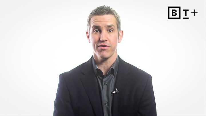 A man in a dark suit jacket and collared shirt speaks in front of a plain white background with a "BT+" logo in the top right corner.