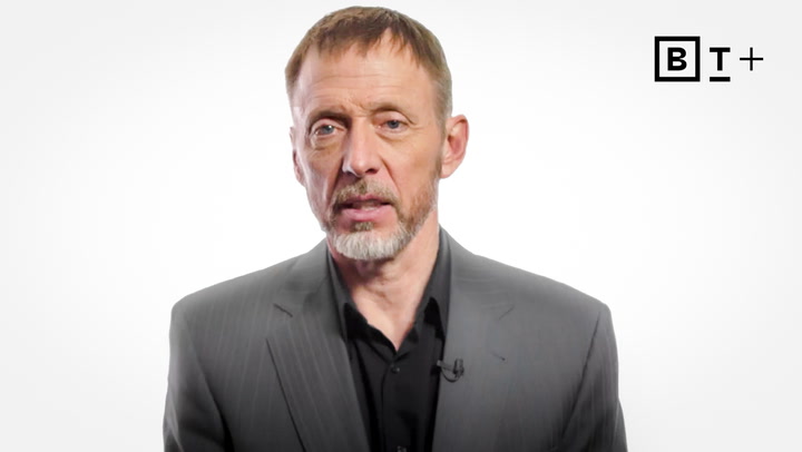A man in a gray suit and black shirt speaks while facing the camera, with a "B T +" logo in the top right corner on a white background.
