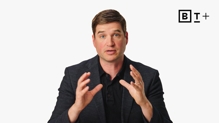 A man in a dark blazer gestures with his hands while speaking, seated in front of a white background with a logo in the top right corner.