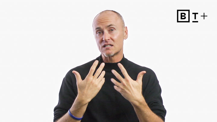 A man in a black long-sleeve shirt gestures with both hands while speaking, against a plain white background with a "B T +" logo in the top right corner.