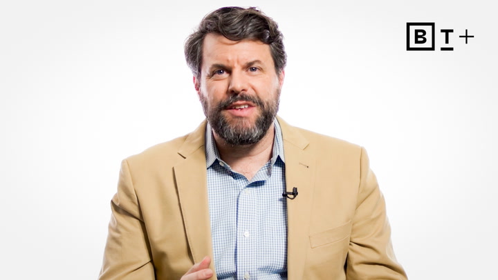A man with a beard and mustache wearing a tan blazer and a light blue checkered shirt speaks in front of a white background with a “B T+” logo in the top right corner.