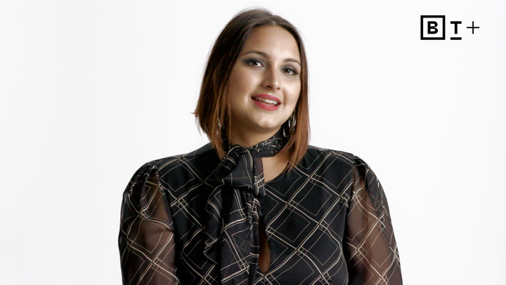 A person with brown hair, wearing a black patterned blouse with a bow, sits in front of a white background with a "BT+" logo in the top right corner.