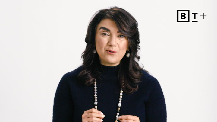 A woman with wavy dark hair wearing a dark sweater and beaded necklace speaks in front of a plain white background with the BT+ logo in the corner.