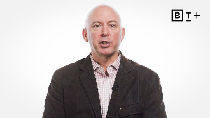 A bald man in a dark jacket and checkered shirt speaks in front of a white background with a "BT+" logo in the top right corner.