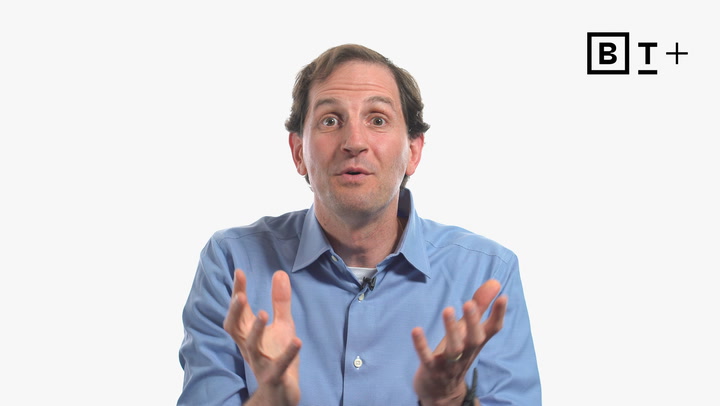A man in a blue shirt gestures with his hands while speaking. A "B T +" logo appears in the top right corner on a plain white background.