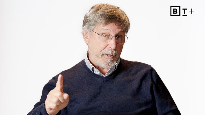 An older man with gray hair and a beard, wearing glasses and a navy sweater, gestures with his index finger raised against a plain white background.