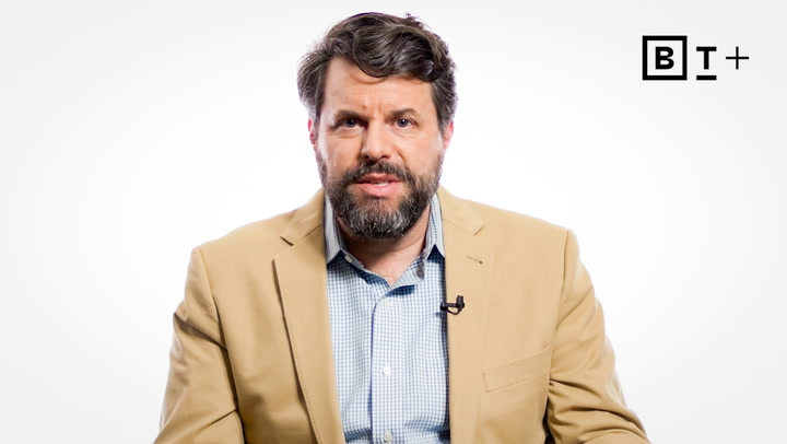 A man with dark hair and a beard, wearing a tan blazer and light blue shirt, sits in front of a white background with a "BT+" logo in the top right corner.