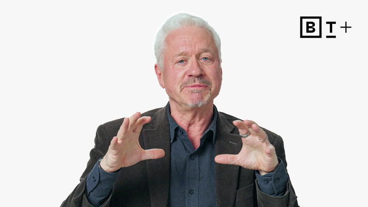 An older man with white hair and a goatee gestures with his hands while speaking; he wears a dark blazer and shirt. A "B T+" logo appears in the top right corner.