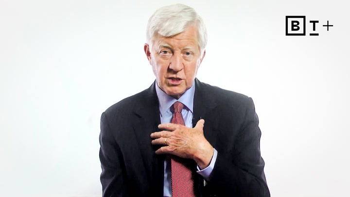 An older man in a suit and red tie speaks while gesturing with his hand on his chest. The background is plain white with a “BT+” logo in the upper right corner.