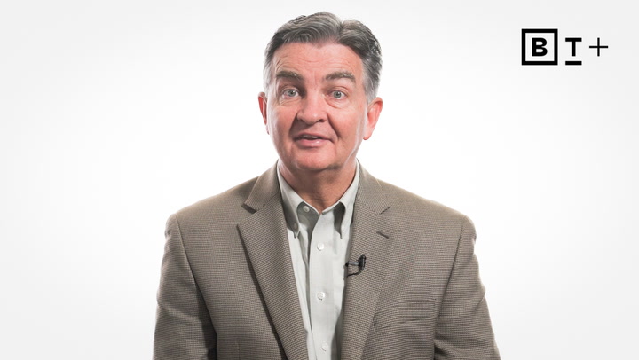 A middle-aged man in a brown blazer and light shirt speaks in front of a white background with the BT+ logo in the top right corner.