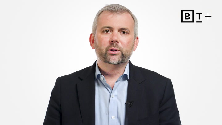 A man in a suit jacket and open-collared shirt speaks in front of a white background with a "B T +" logo in the top right corner.
