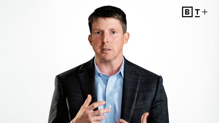 A man in a dark suit and light blue shirt speaks, gesturing with his hand, against a plain white background with the BT+ logo in the top right corner.