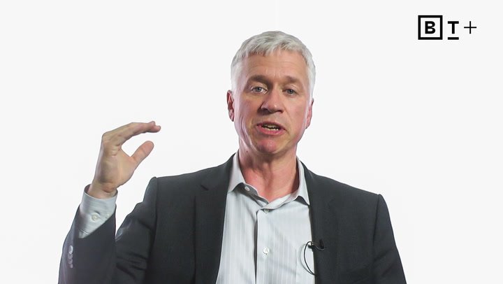 A man in a suit gestures with his right hand raised, while speaking. The background is plain white with a "BT+" logo in the top right corner.