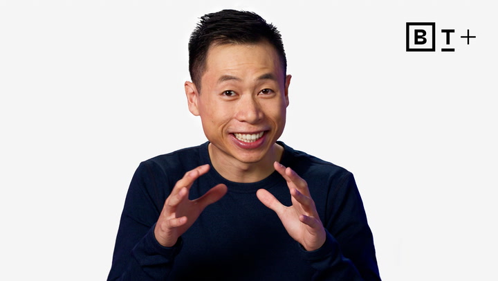 A man in a navy shirt gestures with both hands while smiling, with a "B T +" logo in the top right corner against a white background.