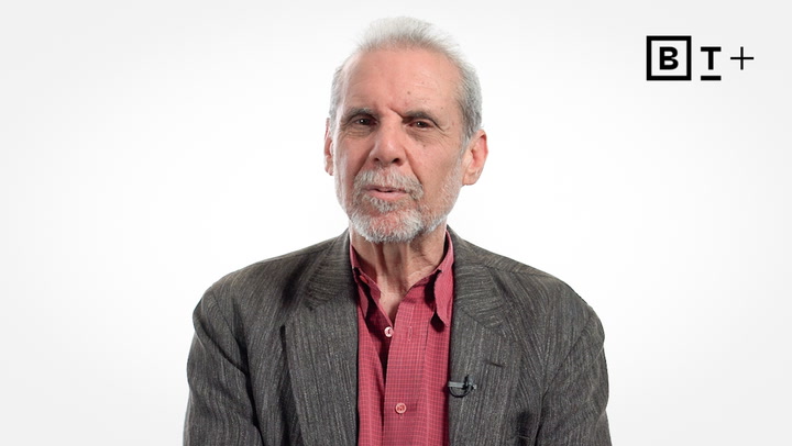 An older man with short gray hair and beard wears a red shirt and gray blazer, seated against a plain white background with a "BT+" logo in the top right corner.
