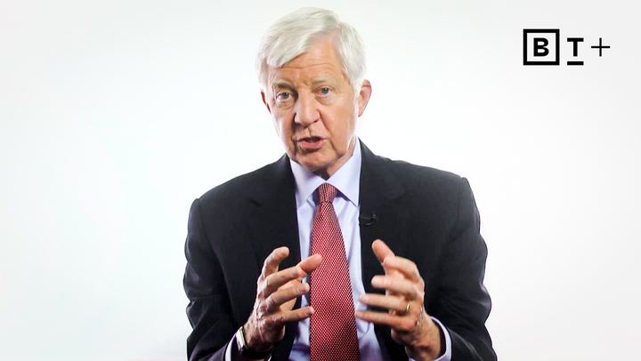 An older man in a dark suit and red tie speaks while gesturing with his hands. There is a "B T +" logo in the top right corner. The background is plain white.