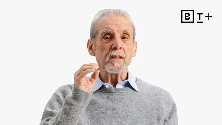 An older man with gray hair and a beard, wearing a gray sweater over a blue collared shirt, gestures with his right hand. A "B T +" logo is in the top right corner on a white background.